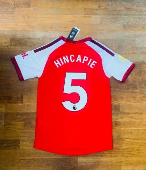 Arsenal tröja Hincapie #5 - Snygg röd och vit Arsenal matchtröja från Adidas med Hincapie och nummer 5 på ryggen. Tröjan har klubbmärke och sponsortryck på bröstet, samt detaljer i svart och gult på ärmarna. Perfekt för fotbollsfans som vill sticka ut på läktaren eller planen.
