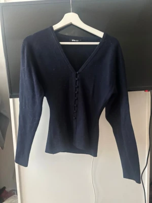 Mörkblå stickad tröja Gina Tricot - Mörkblå stickad tröja från Gina Tricot med v-ringning och dekorativa knappar framtill. Tröjan har långa ärmar och en smal, figurnära passform. Perfekt för dig som gillar stilrena och enkla plagg.