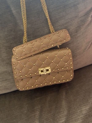 Beige handväska med nitar - Säljer en beige quiltad handväska från V med guldfärgade nitar och kedjerem. Väskan har ett elegant spänne framtill och en röd innerficka. Perfekt accessoar för dig som gillar lyxig och edgy stil. Har lite repor på låset som ni kan se på sista bilden.