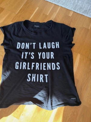 Svart t-shirt med texttryck medium - Svart t-shirt med vit text: 'DON'T LAUGH IT'S YOUR GIRLFRIENDS SHIRT'. Klassisk rund hals och korta ärmar. Perfekt för dig som gillar statement-plagg och vill ha en chill och avslappnad stil.