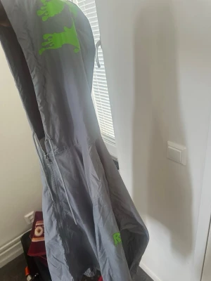 Grå vindjacka med grön detalj - Säljer en grå vindjacka med huva och dragkedja framtill. Jackan har en grön grafisk detalj på huvan och nedtill. Tillverkad i lätt och tunt material, perfekt för blåsiga dagar. Justerbar nederkant och resår vid ärmslut för extra komfort.