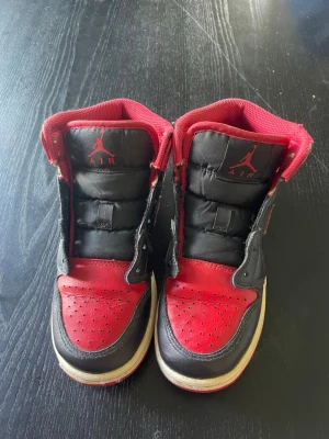 Svarta och röda Air Jordan 1 High - Säljer ett par klassiska Air Jordan 1 High sneakers i svart och rött med vita sulor. Skorna har den ikoniska Nike Swoosh-loggan på sidan och Jumpman-loggan på plösen. Ovandelen är i läder och designen är tidlös och sportig. Priset kan diskuteras 