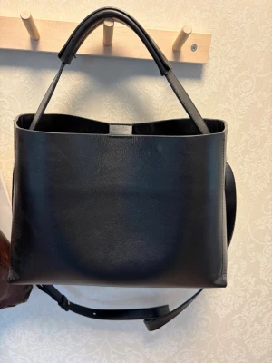 Svart stilren handväska i läder - Snygg svart handväska i slätt läder med minimalistisk design. Väskan har två korta handtag och en avtagbar axelrem för olika bärstilar. Rektangulär form och enkel look som passar till många outfits. Perfekt för dig som gillar klassiska accessoarer.