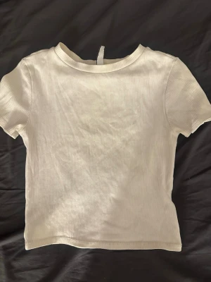 Vit ribbad t-shirt från Divided - En enkel och stilren vit ribbad t-shirt från Divided. T-shirten har korta ärmar, rund halsringning och är gjord i ett mjukt bomullsmaterial. Perfekt basplagg till garderoben och lätt att matcha med allt.