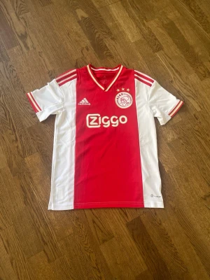 Ajax fotbollströja - Ajax hemmatröja från Adidas i klassisk röd och vit design med klubbmärke och Ziggo-logga på bröstet. Tröjan har korta ärmar, V-ringad krage med röda och vita detaljer samt tre ränder på axlarna. Perfekt för fotbollsträning eller match.