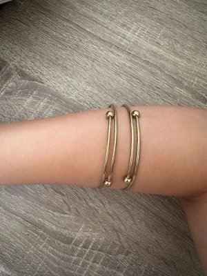 Guldiga minimalistiska armband - Två stilrena armband i guldfärgad metall med minimalistisk design. Armbanden har raka, tunna band och avslutas med små runda kulor på varje ände. Perfekta för dig som gillar enkel men snygg accessoar.