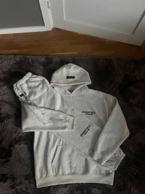 Grå Essentials Fear of God set - Säljer ett grått loungewear-set från Essentials Fear of God. Setet består av en hoodie med svart logga på bröstet och matchande mjukisbyxor med dragsko och resår i midjan. Båda plaggen är i mjuk bomullsblandning och har en avslappnad passform.