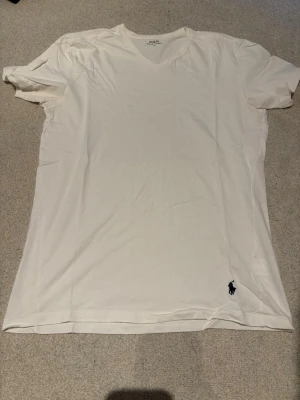 Vit t-shirt med v-ringning Polo Ralph Lauren - Klassisk vit t-shirt från Polo Ralph Lauren med v-ringning och korta ärmar. Tillverkad i mjukt bomullsmaterial och har den ikoniska mörkblå logotypen broderad nertill på vänster sida. Perfekt basplagg för en clean och stilren look.