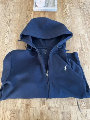 Marinblå zip hoodie från Polo Ralph Lauren - Snygg marinblå hoodie från Polo Ralph Lauren med dragkedja framtill och klassisk huva. Tröjan har det ikoniska vita Polo-logotrycket på bröstet och är tillverkad i mjukt bomullsmaterial. 