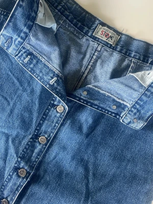 Lång jeanskjol med knappar - Säljer en klassisk blå lång jeanskjol med knappar hela vägen framtill. Kjolen har en rak passform och är tillverkad i kraftigt denimtyg. Perfekt för dig som gillar en tidlös och avslappnad stil.