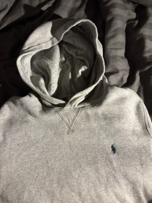 Grå hoodie från Polo Ralph Lauren - Snygg grå hoodie från Polo Ralph Lauren med klassisk huva och den ikoniska blå loggan broderad på bröstet. Tillverkad i mjukt sweatshirtmaterial och har en känguruficka framtill. Perfekt för en chill och avslappnad stil.