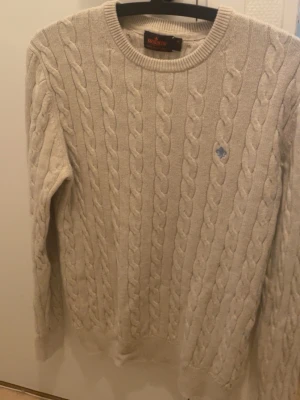 Beige kabelstickad tröja Morris M - Kabelstickad tröja från Morris i beige färg, tillverkad av 100% pima bomull. Rund halsringning, ribbade muddar och diskret blå logga broderad på bröstet. Perfekt för dig som gillar klassisk och stilren look.