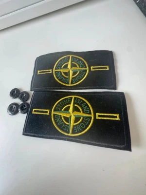 Stone Island tygmärken med knappar - Två svarta tygmärken från Stone Island med det klassiska gula och gröna kompasslogotypen, samt fyra svarta knappar. Perfekt för att sy eller fästa på jackor, tröjor eller väskor för en unik look.