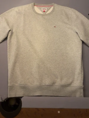 Grå sweatshirt från Tommy Hilfiger - Snygg grå sweatshirt från Tommy Hilfiger med rund halsringning och diskret broderad logga på bröstet. Tröjan har ribbade muddar vid ärmslut och nederkant samt klassisk passform. Perfekt för en avslappnad och stilren look.