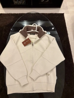 Vit ziptröja med brun krage Loro piana  - Snygg vit ziptröja med ribbstickade muddar och bred brun krage. Tröjan har hel dragkedja framtill och är gjord i ett tjockare stickat material. Perfekt för lager på lager och ger en clean look. Loro piana