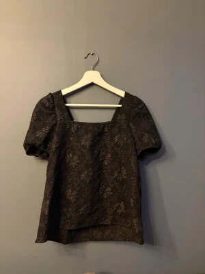 Svart blus med puffärm och rosett - Snygg svart blus med diskret blommönster, puffiga korta ärmar och fyrkantig ringning. Baksidan har en vit rosett som detalj. 