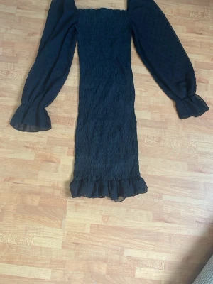 Svart långärmad klänning med volang - Svart klänning med smockad kropp och transparenta långärmade ärmar med diskret prickigt mönster. Volangdetaljer vid ärmslut och nederkant. 