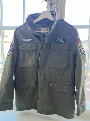 Cotton Herringbone Field Jacket - kille  - Snygg militärgrön vindjacka från Polo Ralph Lauren med patchar på ärmarna och bröstet. Jackan har fyra fickor framtill, hög krage och dragkedja. Materialet är slitstarkt bomullstyg. 