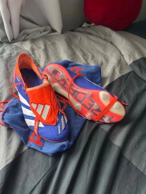 Adidas Predator blå/orange fotbollsskor - Säljer ett par Adidas Predator fotbollsskor i blått och orange med vita detaljer. Skorna har en modern design med snörning, låg profil och mönstrad sula för bra grepp på planen. Tillverkade i syntetmaterial och levereras med en blå meshpåse.