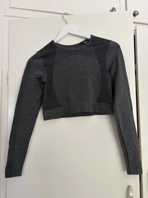 Mörkgrå croppad träningströja - Snygg mörkgrå croppad träningströja med långa ärmar och rund hals. Tillverkad i stretchigt funktionsmaterial som andas, perfekt för gymmet eller löprundan. Tröjan har diskreta mönsterdetaljer och sitter tight för en sportig look.