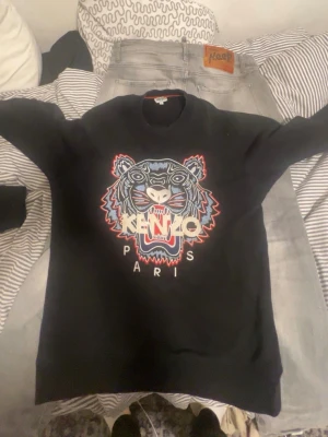 Svart sweatshirt från Kenzo med tiger - Svart sweatshirt från Kenzo med ikoniskt tigerbroderi i blått, rött och vitt på bröstet samt texten 'KENZO PARIS'. Rund halsringning och mjukt material som känns skönt mot huden. Perfekt för dig som gillar streetwear och vill sticka ut.
