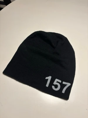Svart mössa med trycket 157 - Säljer en svart mössa i mjukt stickat material med siffran 157 tryckt i vitt framtill. Enkel och stilren design som passar till det mesta. Perfekt accessoar för kalla dagar.