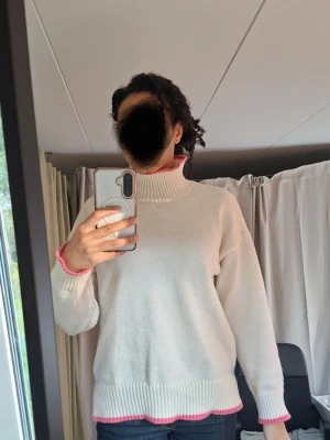 Vit polotröja med rosa detaljer - Säljer en krämvit stickad polotröja med ribbad hög krage och snygga rosa detaljer vid ärmslut och nederkant. Tröjan har en avslappnad passform och är perfekt för lager på lager. Materialet ser mjukt och bekvämt ut, och modellen är långärmad.
