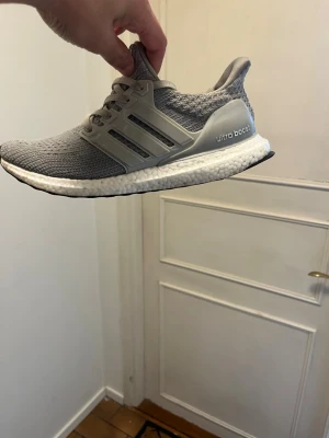 Grå Adidas Ultra Boost sneakers - Säljer ett par grå Adidas Ultra Boost sneakers med stickad ovandel och vita Boost-sulor. Skorna har klassiska tre ränder på sidan och en bekväm, sportig design. Perfekta för dig som gillar stilrena och bekväma sneakers.