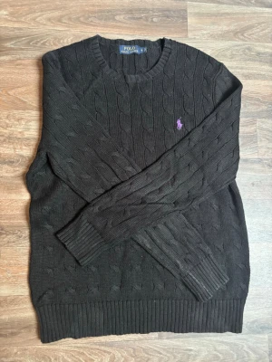 Svart kabelstickad tröja Polo Ralph Lauren - Kabelstickad svart tröja från Polo Ralph Lauren med rund halsringning och lila broderad logga på bröstet. Tröjan har ribbade muddar vid ärmslut och nederkant, och är tillverkad i ett mjukt stickat material som passar perfekt till höst och vinter.