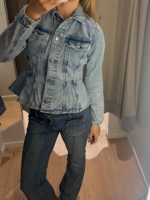 Ljusblå figursydd jeansjacka - Trendig ljusblå jeansjacka med figursydd passform och markerad midja. Klassisk krage, två bröstfickor med lock och silverfärgade knappar framtill. Snygga sömdetaljer ger jackan en modern look. Perfekt att styla till jeans eller kjol.