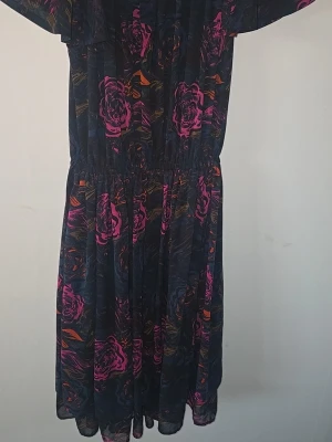 Mörkblommig klänning från H&M - Snygg mörkblommig klänning från H&M i storlek 34. Klänningen har ett svart basmaterial med rosa och lila blommönster samt inslag av orange. Den har resår i midjan och ett luftigt, lätt tyg som ger en avslappnad siluett.