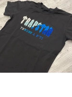 Svart Trapstar t-shirt med brodyr - Säljer en svart t-shirt med broderad Trapstar-text i vitt och blått på bröstet. T-shirten har klassisk passform och korta ärmar. Materialet känns som bomull och trycket har en cool struktur som sticker ut.