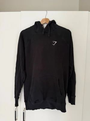 Svart hoodie från gymshark - Säljer en svart hoodie från gymshark med vit logga på bröstet. Hoodien har huva med dragsko, stor magficka och ribbade muddar vid ärmar och nederkant.  Storlek S men passar även för M. Bra skick.