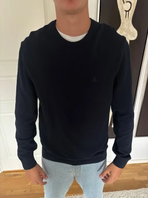 Mörkblå stickad tröja Marc O'Polo - Snygg mörkblå stickad tröja från Marc O'Polo med rund halsringning och diskret struktur i materialet. Perfekt för lager-på-lager och enkel att matcha med jeans eller chinos. Klassisk modell med ribbade muddar vid ärmslut och nederkant.
