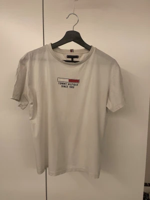 Vit t-shirt från Tommy Hilfiger - Snygg vit t-shirt från Tommy Hilfiger med tryck på bröstet där det står 'Tommy Hilfiger Since 1985'. Klassisk rund halsringning och korta ärmar. Tillverkad i mjuk bomull som känns skön mot huden. Perfekt till jeans eller shorts.