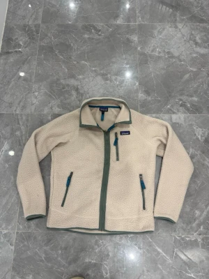 Beige fleecejacka från Patagonia - Säljer en beige fleecejacka från Patagonia med gröna detaljer och dragkedjor. Jackan har tre fickor med blå dragkedjor och hög krage. Perfekt för lager-på-lager och outdoor-vibe. Klassisk logga på bröstet och mjukt, mysigt material.