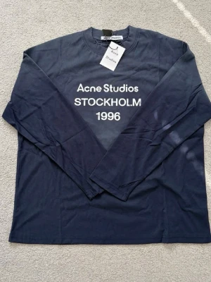 Acne Studios longsleeve  - Snygg mörkblå långärmad topp från Acne Studios med vit text framtill: 'Acne Studios STOCKHOLM 1996'. Klassisk rund halsringning och relaxed fit. Perfekt för dig som gillar stilrena och trendiga plagg.