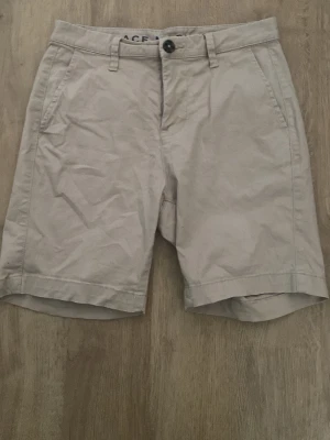 Beige chinoshorts med fickor - Snygga beige chinoshorts med klassisk passform och bälteshällor. Shortsen har två sidofickor fram och två passpoalfickor bak. Tillverkade i ett mjukt bomullstyg som är perfekt för varma dagar. Stängs med knapp och dragkedja.