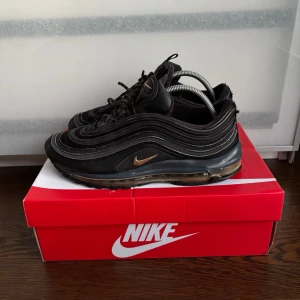 Nike Air Max 97 svarta sneakers - Nike Air Max 97 sneakers i svart med guldfärgade detaljer och synlig Air-sula. Skorna har en vågig design i syntet och mesh, samt svarta skosnören. Perfekta för dig som gillar en sportig och stilren look. 42,5