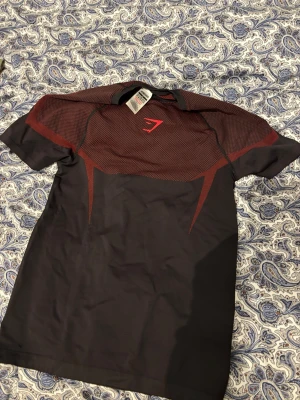 Svart och röd träningströja onyx Gymshark - Snygg svart och röd träningströja från Gymshark med korta ärmar och sportig design. Tröjan har ett mönstrat parti över bröstet och axlarna samt Gymshark-logga framtill. Tillverkad i ett lätt och stretchigt material som passar perfekt för gymmet.