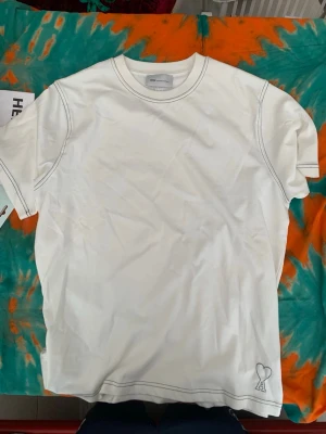 Vit t-shirt från Ami med hjärtdetalj - Säljer en stilren vit t-shirt från Ami Alexandre Mattiussi med svart kontrastsöm och ett litet hjärta broderat nere vid fållen. Klassisk passform och rund halsringning. Perfekt till jeans eller shorts.