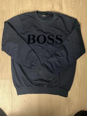 Mörkblå stickad tröja från Hugo Boss - Mörkblå stickad tröja från Hugo Boss med stor BOSS-logga framtill. Tröjan har rund halsringning, ribbade muddar vid ärmslut och nederkant samt en klassisk, avslappnad passform. Perfekt för dig som gillar stilrena och tidlösa plagg.