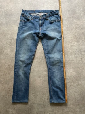 Nudie jeans - Säljer ett par klassiska blå jeans med raka ben och kontrastsömmar. Jeansen har fem fickor, knappgylf och subtila slitningar framtill. Materialet är denim i bomull och passformen är normal. Perfekta till en avslappnad stil.