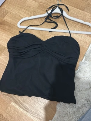 Svart halterneck linnetopp från NELLY - Snygg svart linnetopp med halterneck och rynkade detaljer över bysten. Toppen är ärmlös och har en croppad passform som ger en trendig look. Perfekt att matcha med jeans eller kjol för en cool stil.