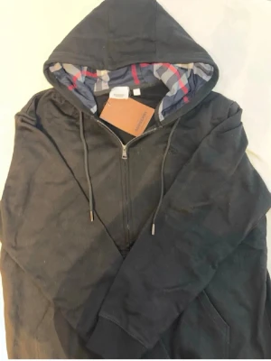 Svart zip hoodie från Burberry - Svart hoodie med dragkedja från Burberry. Klassisk huva med dragsko och insida i Burberrys rutiga mönster i grått, rött och vitt. Metalländar på snörena och detaljer med Burberry-logga. Perfekt för en stilren och avslappnad look.