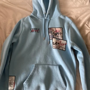 Ljusblå hoodie från Supply & Demand - Säljer en ljusblå hoodie från Supply & Demand med tryck av palmer och textdetaljer. Hoodien har en stor ficka framtill och justerbar huva. Perfekt för en avslappnad stil. 175-185