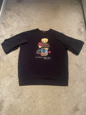 Svart sweatshirt med Polo Bear - TRÖJAN ÄR XL I BARN STORLEK.          Cool mörkblå sweatshirt från Ralph Lauren med den ikoniska Polo Bear-björnen på bröstet. Björnen har på sig en stickad tröja och jeans, och trycket är färgglatt och detaljerat. Tröjan har rund hals och avslappnad passform, perfekt för en casual look.