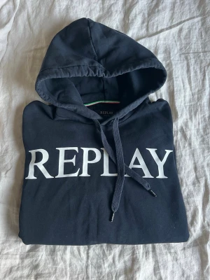 Blå hoodie från Replay - Cool blå hoodie från Replay med stor vit logotyp på bröstet och matchande blåa snören med Replay-detaljer. Klassisk huva och mjukt material som känns skönt mot huden. Perfekt för dig som gillar stilrent. Storlek L men passar L/M.