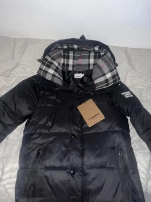 Svart pufferjacka från Burberry - Snygg svart pufferjacka från Burberry med klassiskt rutigt foder i huvan och tydlig Burberry London England-patch på ärmen. Jackan har dragkedja och knappar framtill samt två fickor med dragkedja. Perfekt för kalla vinterdagar. Går it göra t väst 
