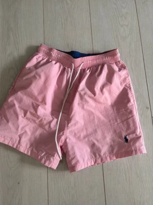 Rosa badbyxor från Polo Ralph Lauren - Snygga rosa badbyxor från Polo Ralph Lauren med elastisk midja och vit dragsko. Klassisk mesh-innerbyxa och blå insida i midjan. Liten mörkblå logga broderad på vänster ben. Perfekta för stranden eller poolen.